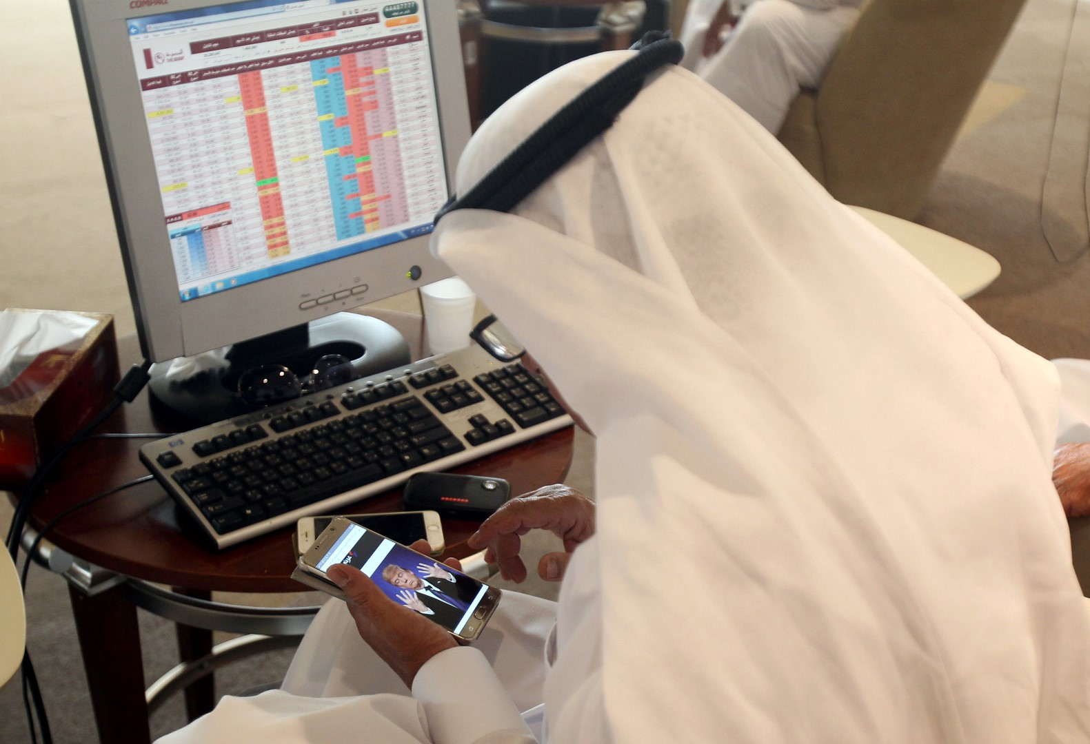 A trader uses his smartphone to follow news of U.S. presidential elections as he monitors a screen displaying stock information at Qatar Stock Exchange in Doha, Qatar November 9, 2016. REUTERS/Naseem Zeitoon - RTX2SQ7Q